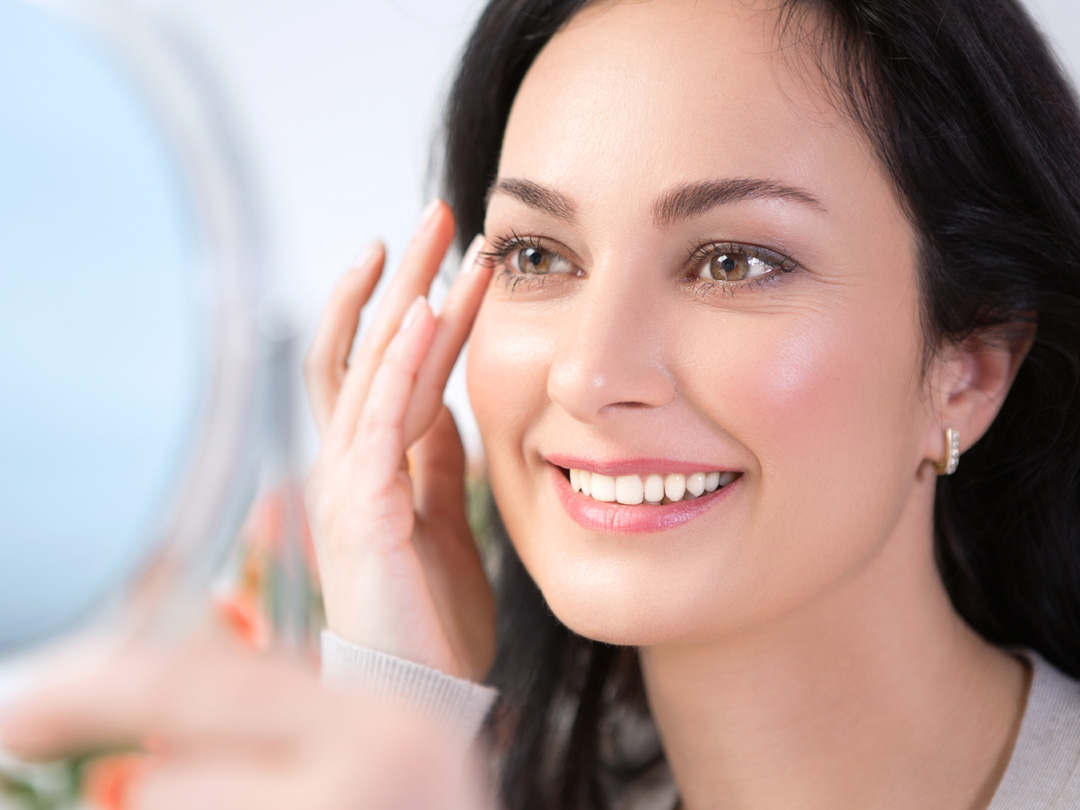 A woman smiling with joy at her reflection while touching her radiant skin, showing the confidence that comes from effective skin support.
